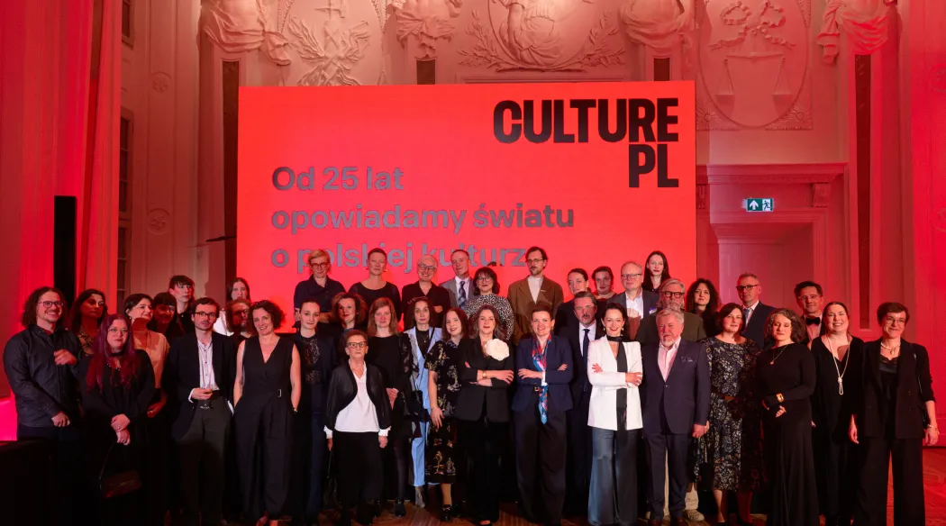 Grupowe zdjęcie kilkudziesięciu osób w eleganckich strojach w sali z rzeźbionymi płaskorzeźbami. Za nimi czerwony ekran z logo Culture.pl i napisem „Od 25 lat opowiadamy światu o polskiej kulturze”.