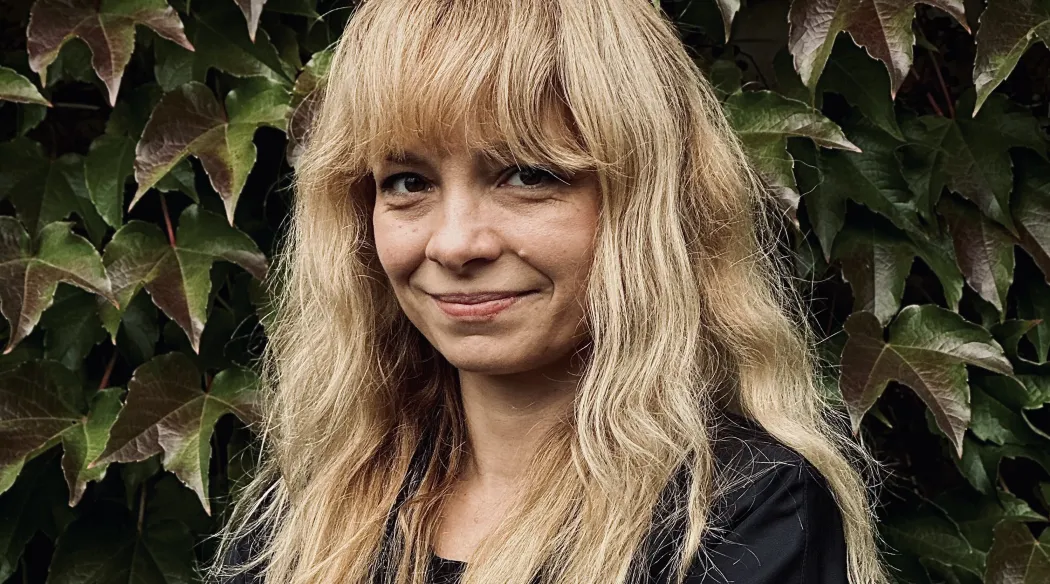 Portrait of Marta Lisok. A smiling woman with long blond hair and a fringe, wearing a black blouse, stands with her arms crossed in front of a wall covered with ivy.