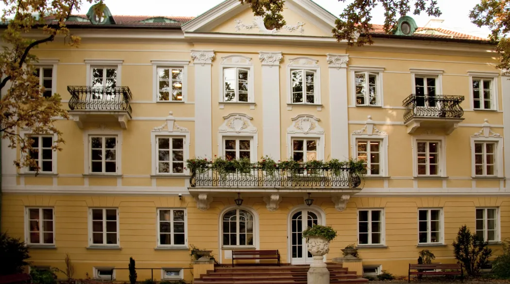 The façade of Pałacyk Cukrowników, the seat of the Adam Mickiewicz Institute: a yellow three-storey building with white ornaments, a triangular pediment and wrought-iron balconies, one of them decorated with flowers. In front of the entrance are steps, benches and a stone urn on a pedestal, with autumn tree branches in the foreground.