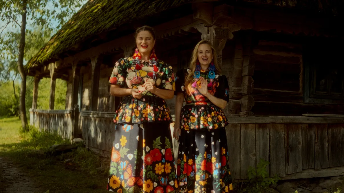 dwie kobiety w ludowych kwiecistych strojach na tle drewnianej chaty