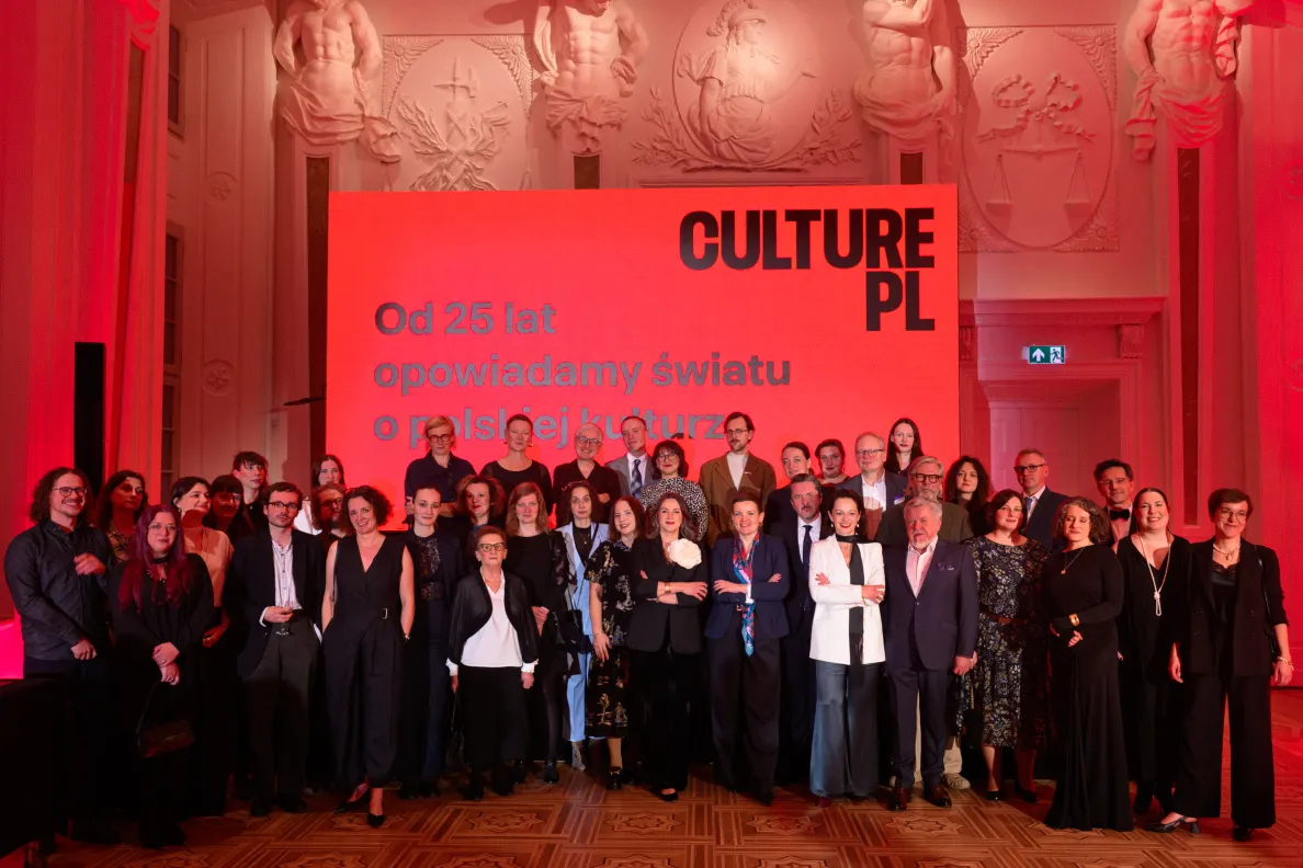 Grupowe zdjęcie kilkudziesięciu osób w eleganckich strojach w sali z rzeźbionymi płaskorzeźbami. Za nimi czerwony ekran z logo Culture.pl i napisem „Od 25 lat opowiadamy światu o polskiej kulturze”.