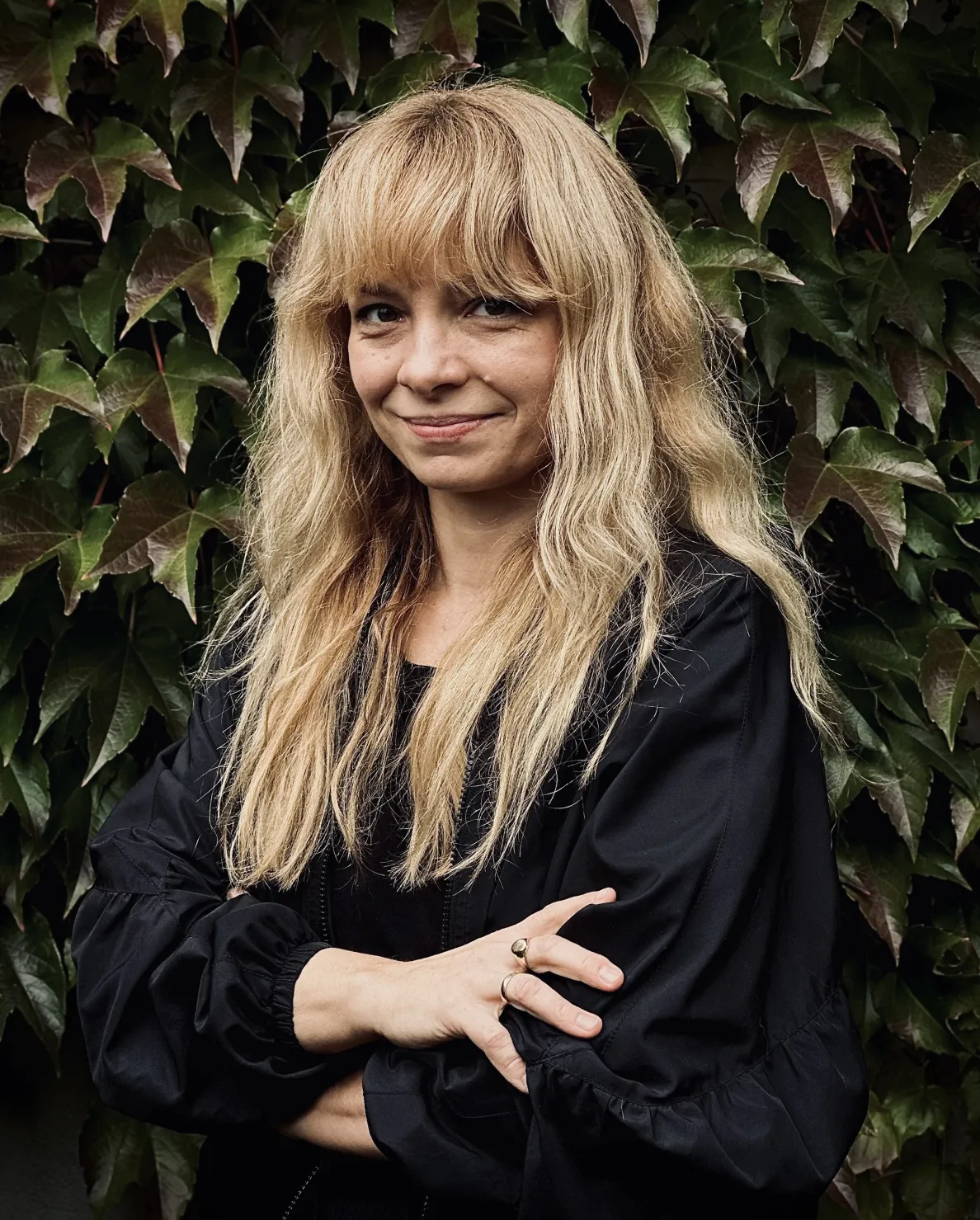 Portrait of Marta Lisok. A smiling woman with long blond hair and a fringe, wearing a black blouse, stands with her arms crossed in front of a wall covered with ivy.