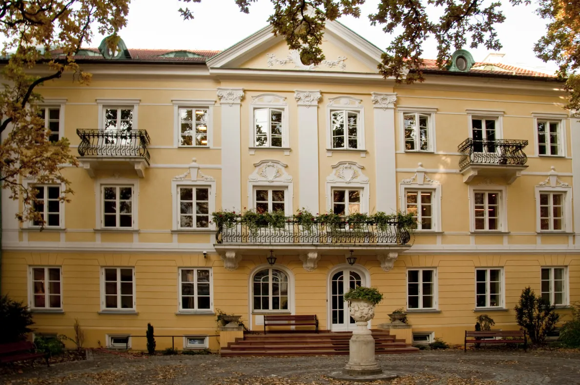 The façade of Pałacyk Cukrowników, the seat of the Adam Mickiewicz Institute: a yellow three-storey building with white ornaments, a triangular pediment and wrought-iron balconies, one of them decorated with flowers. In front of the entrance are steps, benches and a stone urn on a pedestal, with autumn tree branches in the foreground.