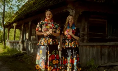 dwie kobiety w ludowych kwiecistych strojach na tle drewnianej chaty