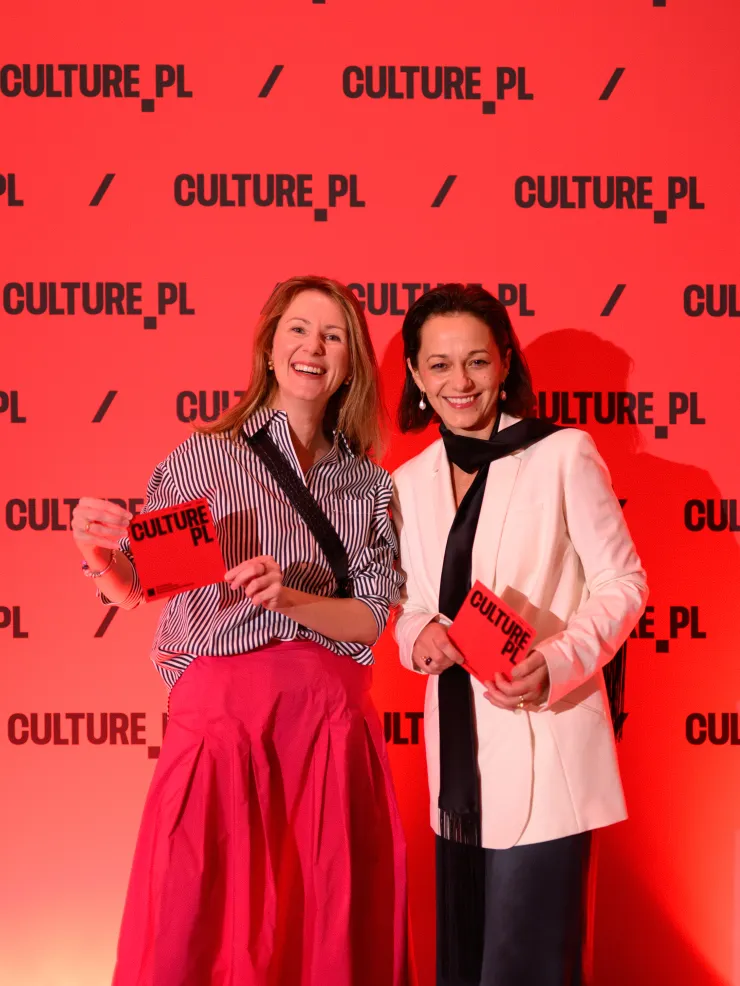 Dwie uśmiechnięte kobiety pozują przed czerwoną ścianką z logo Culture.pl, trzymając czerwone broszury Culture.pl.