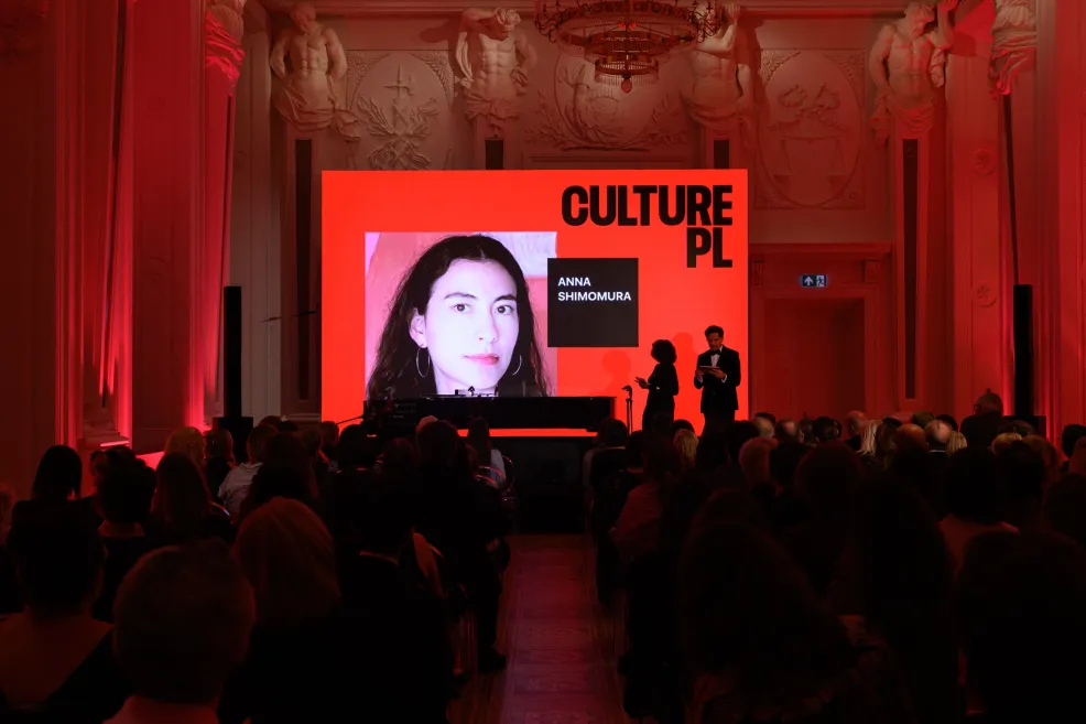 Widok sali z publicznością od tyłu. Na scenie dwoje prowadzących, za nimi czerwony ekran z logo Culture.pl i zdjęciem portretowym podpisanym „Anna Shimomura”.