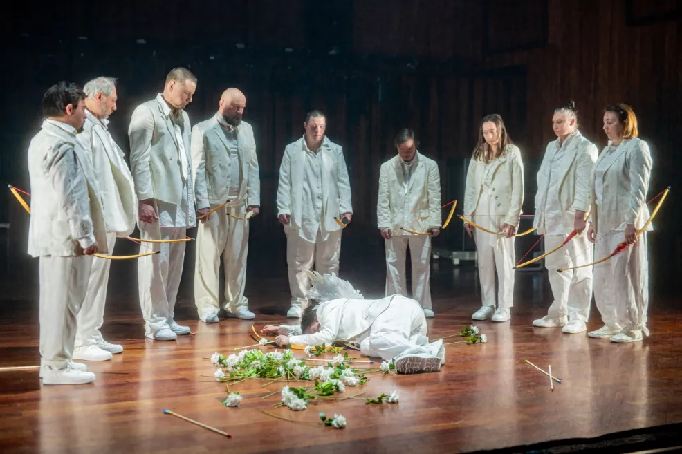 Aktorzy na scenie w półkolu trzymający łuki, na środku leży postać trafiona strzałami
