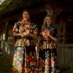 dwie kobiety w ludowych kwiecistych strojach na tle drewnianej chaty