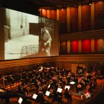 Sala koncertowa, grająca orkiestra i ekran z wyświetlonym kadrem z filmu Wajdy