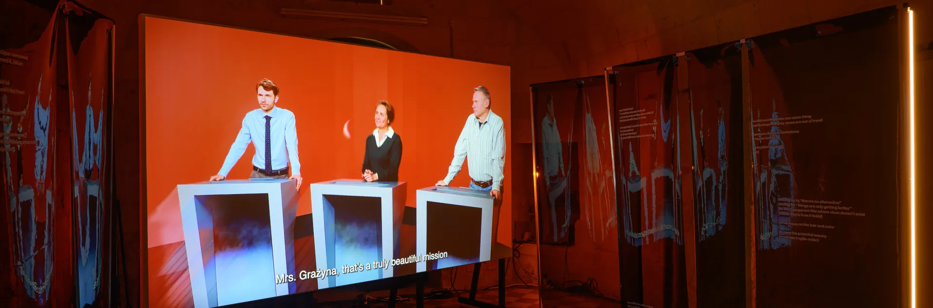 Instalacja Archiwum wahań, trzy osoby biorące udział w teleturnieju widoczne na ekranie ustawionym w pomieszczeniu