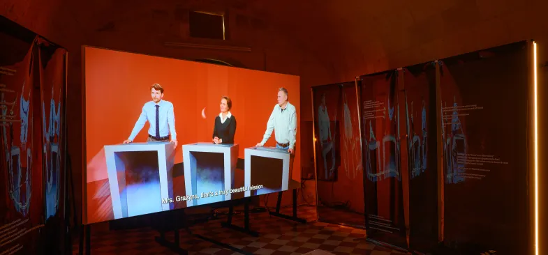 Instalacja Archiwum wahań, trzy osoby biorące udział w teleturnieju widoczne na ekranie ustawionym w pomieszczeniu