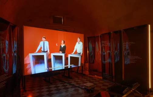 Instalacja Archiwum wahań, trzy osoby biorące udział w teleturnieju widoczne na ekranie ustawionym w pomieszczeniu