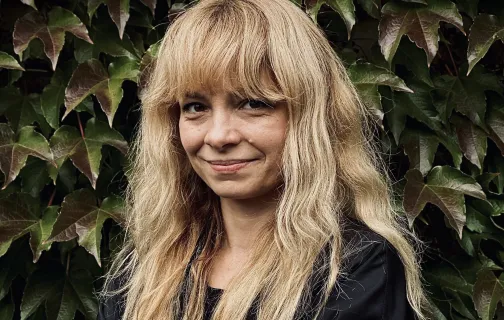 Portret Marty Lisok. Uśmiechnięta kobieta z długimi jasnymi włosami i grzywką, w czarnej bluzce, stoi ze skrzyżowanymi rękami na tle ściany porośniętej bluszczem.